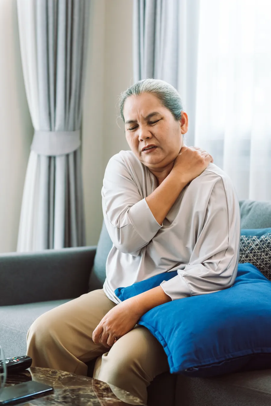 Mature woman sitting at home holding her shoulder in pain, representing the chronic shoulder pain evaluated for Shoulder TAME