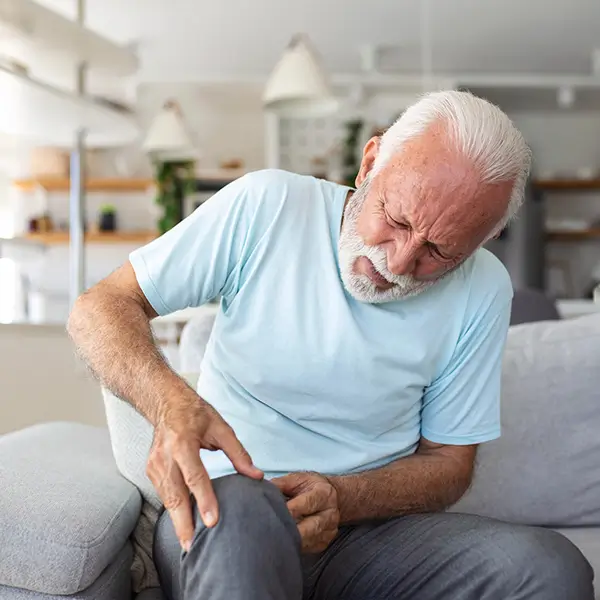 Older man sitting on a couch holding his knee in pain, demonstrating symptoms commonly treated with Genicular Artery Embolization